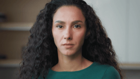 Woman with long curly hair wearing a green top looking directly at the camera with a neutral expression.